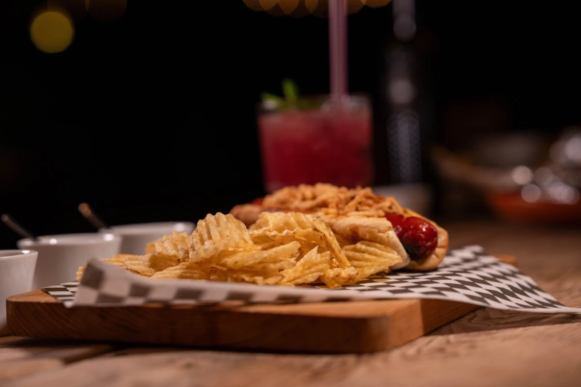 Hot dog with potato chips on a serving board with a red drink