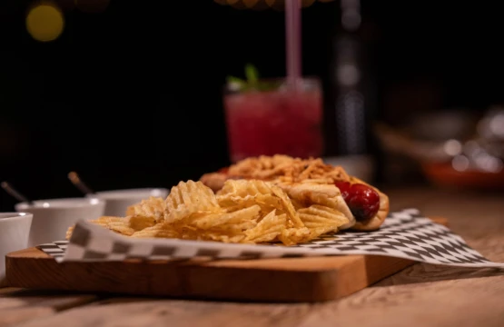 Hot dog with potato chips on a serving board with a red drink